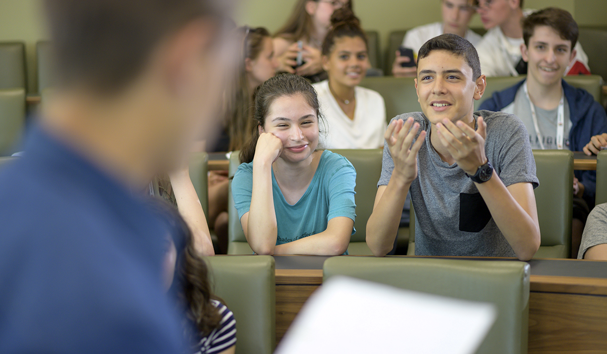 A student gesticulates with his hands during a debate while other students in the audience laugh and smile.