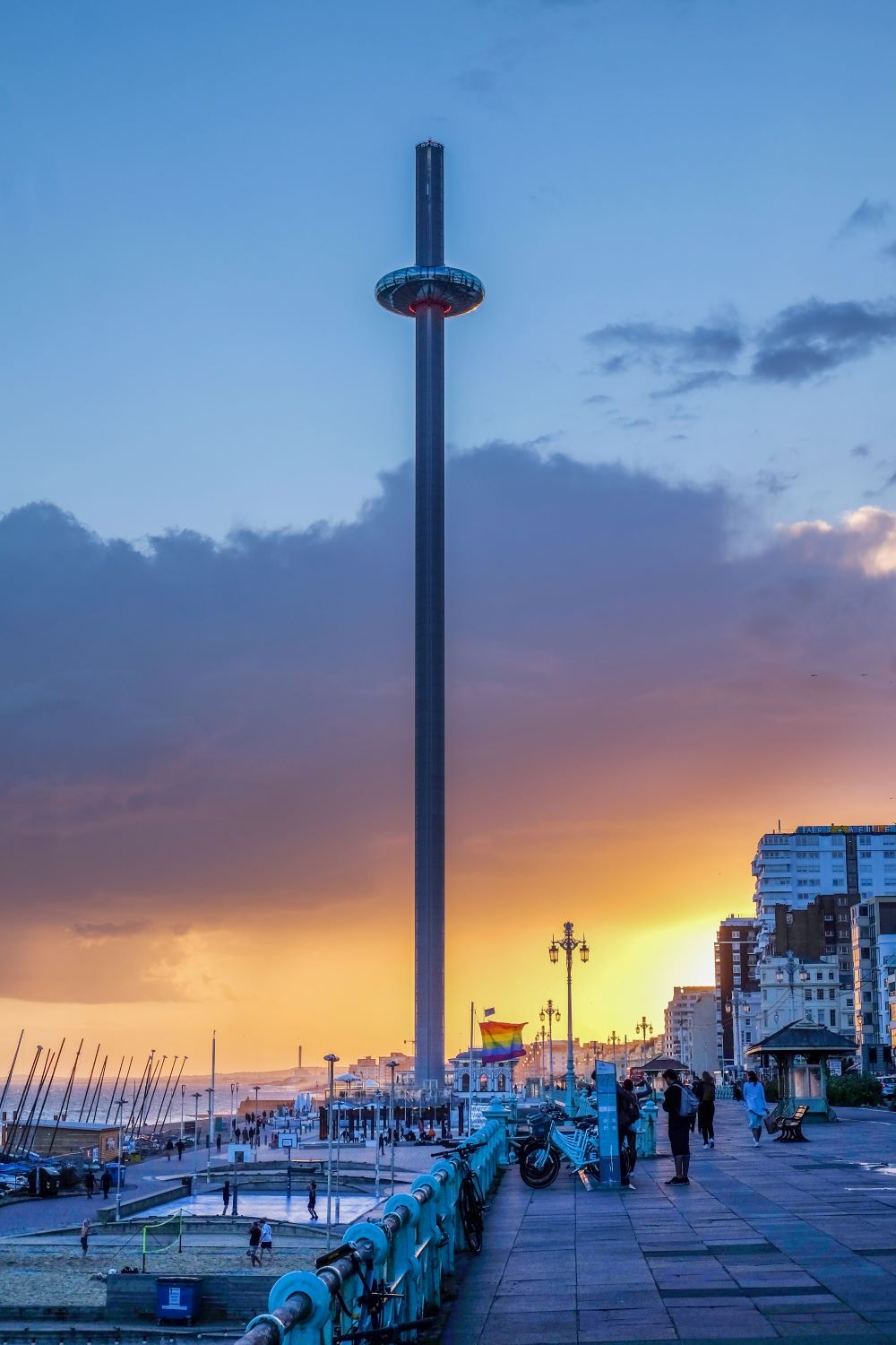 A vivid photo of the i360, where the viewing platform is almost at the top of the vast tower it travels up. Below you can see the famous turquoise railings on the seafront, a rainbow flag and the setting sun glowing in the background.